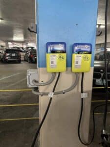 Two Harbor electric vehicle charging stations mounted on a blue pillar inside a parking garage, with several parked cars in the background.