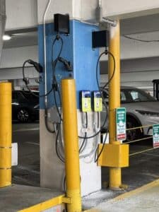 Two electric vehicle charging stations mounted on a blue and white pillar in a parking garage, with yellow protective posts and parked cars nearby.