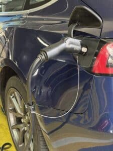 A blue electric car is plugged into a charging station via a charging cable connected to its rear left side.