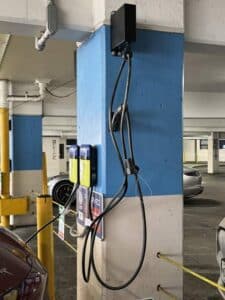 Electric vehicle charging station mounted on a blue pillar in a parking garage, with charging cables hanging down and cars parked nearby.