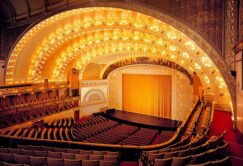 Ornate theater interior with a large, arched ceiling illuminated by rows of lights, red seats, and a golden curtain on stage.