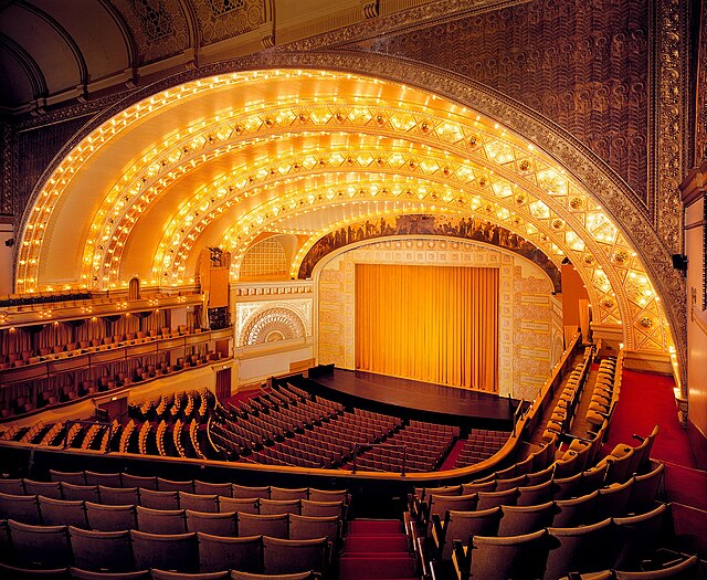 Ornate theater interior with a large, arched ceiling illuminated by rows of lights, red seats, and a golden curtain on stage.