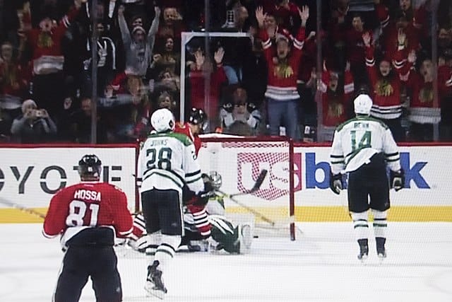 A hockey player scores a goal as fans celebrate in the stands; players from both teams are visible near the net.