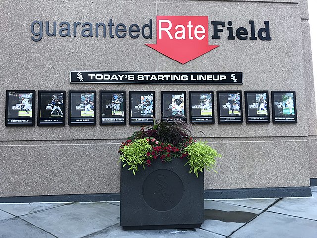 Guaranteed Rate Field wall with starting lineup display boards for a Chicago White Sox game; a planter with flowers sits in front of the display.