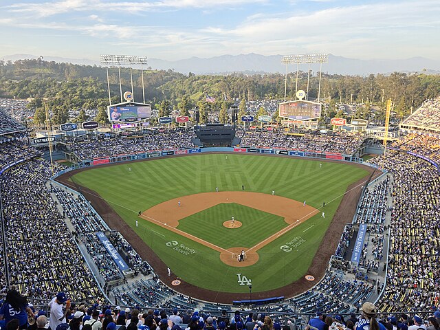 Aerial view of a baseball game in progress at Dodger Stadium, with a full crowd in the stands and the field set against a backdrop of hills and mountains.