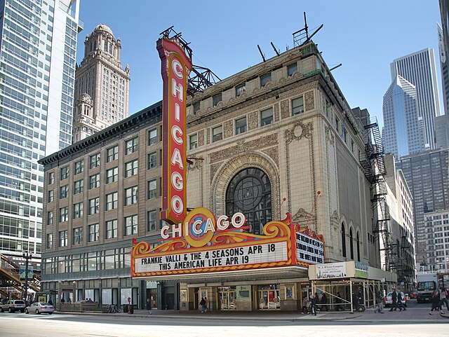 Historic Chicago Theatre with its iconic vertical sign and marquee, surrounded by tall city buildings, under a clear blue sky.