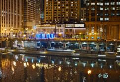 Night view of city buildings and lit restaurants along a riverside, with reflections of lights visible on the water in the foreground.