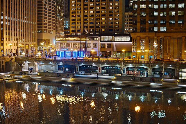 Night view of city buildings and lit restaurants along a riverside, with reflections of lights visible on the water in the foreground.