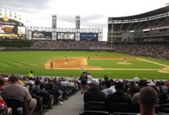 View from the stands of a baseball game at a stadium, showing players on the field, fans in the seats, and a scoreboard in the background.