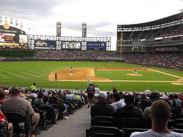 View from the stands of a baseball game at a stadium, showing players on the field, fans in the seats, and a scoreboard in the background.