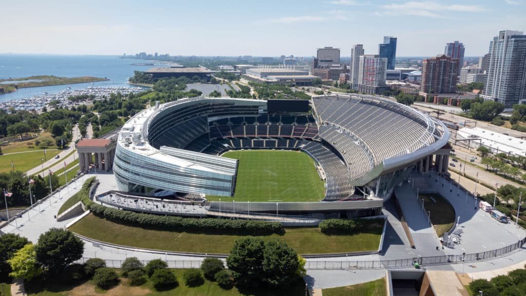 Aerial view of Soldier Field stadium with an empty green field, surrounded by city buildings, trees, and Lake Michigan in the background.