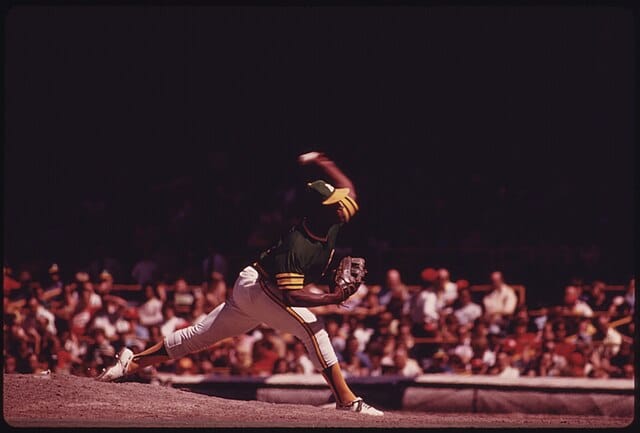 A baseball pitcher in a green and yellow uniform throws a pitch on a mound during a game, with a crowd of spectators in the background.