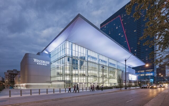 Exterior view of Wintrust Arena at dusk, showing its modern glass facade and people walking on the sidewalk in front of the building.