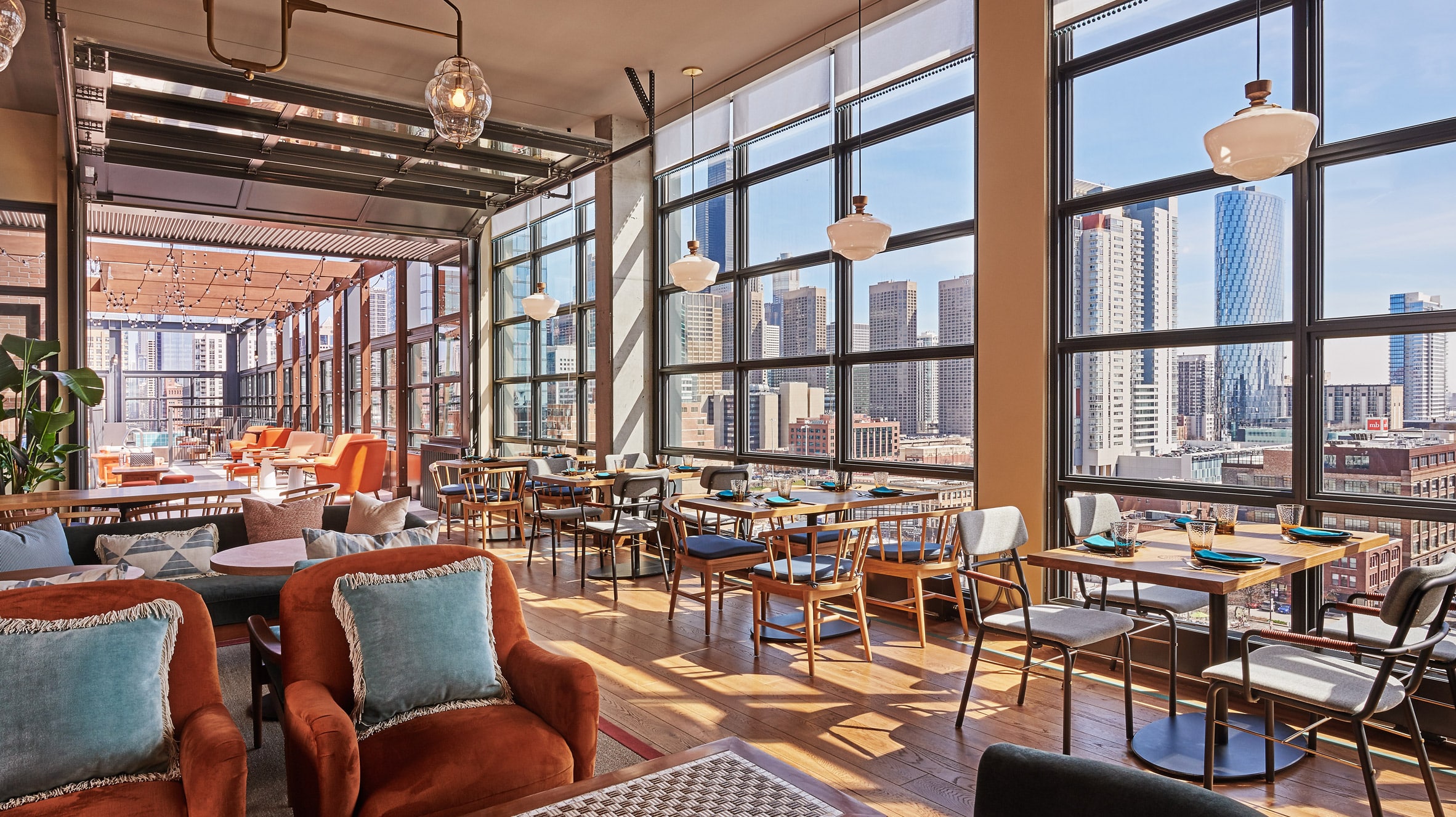 Modern restaurant interior with large windows, city skyline views, wooden floors, tables set for dining, upholstered chairs, and natural daylight streaming in.