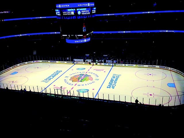An empty ice hockey rink with Chicago Blackhawks logo at center, playoff markings on the ice, and a scoreboard overhead in a large arena.