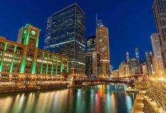 Night view of downtown Chicago with illuminated buildings, including a clock tower, reflected in the Chicago River under a clear sky.