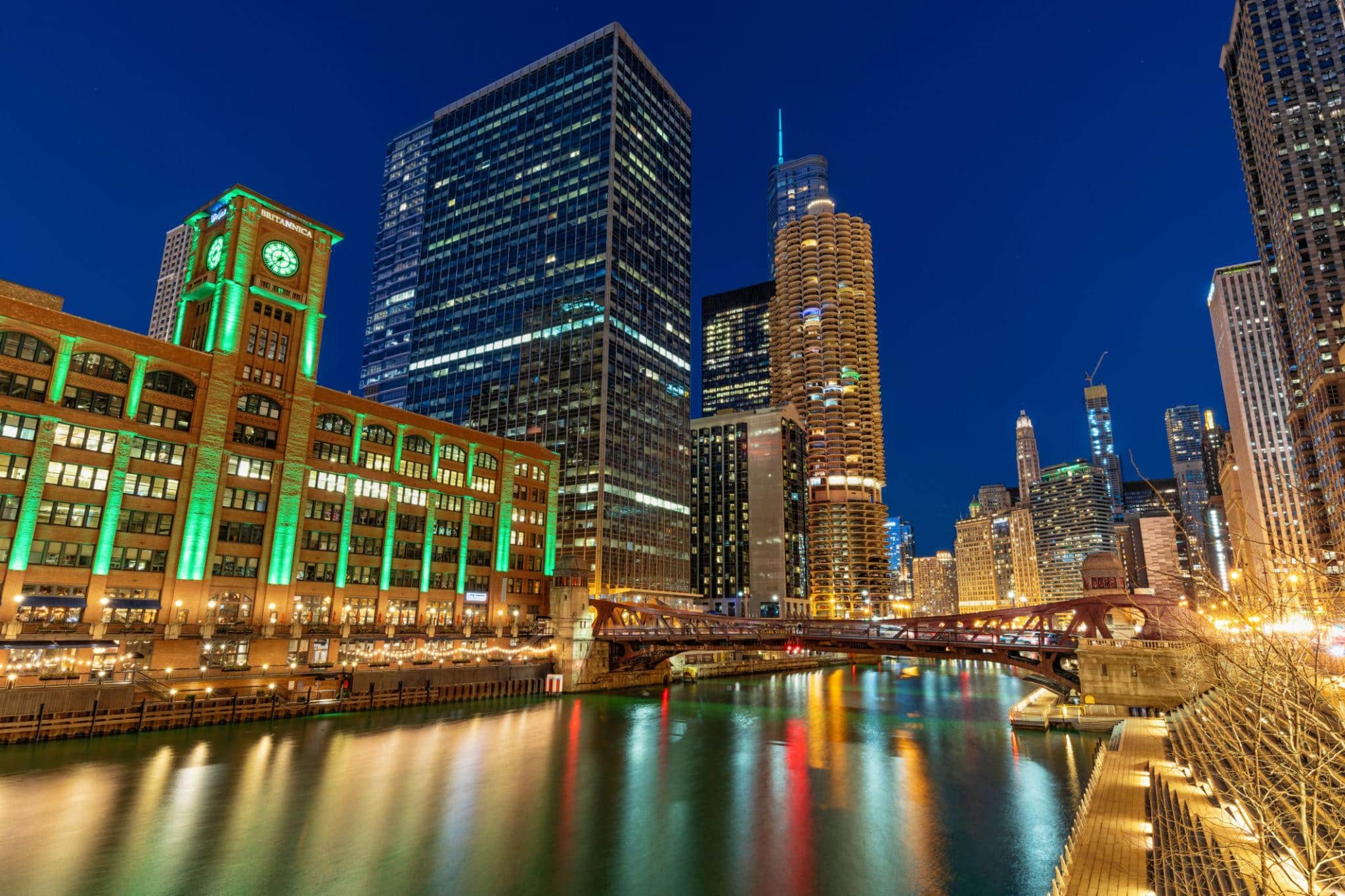 Night view of downtown Chicago with illuminated buildings, including a clock tower, reflected in the Chicago River under a clear sky.