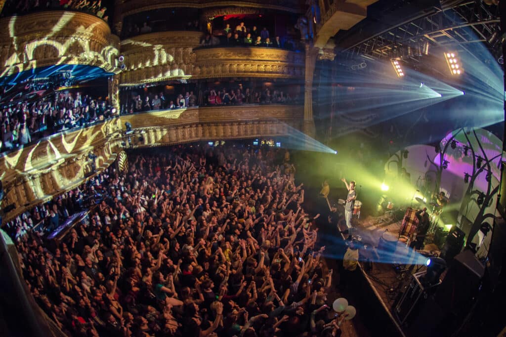 A large crowd watches a live band perform on stage in a theater with ornate balconies and dramatic lighting effects.