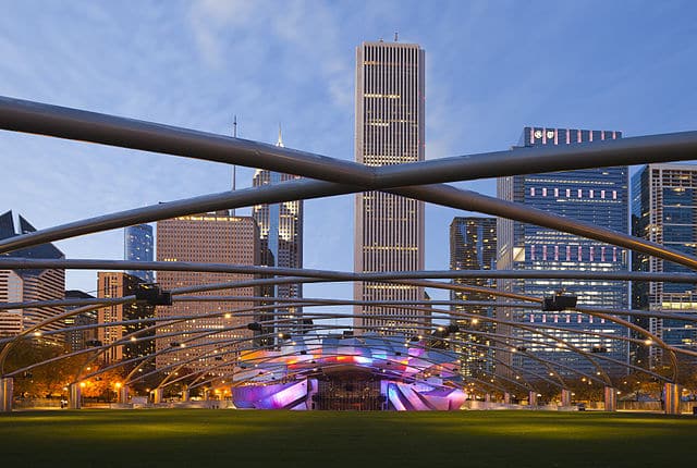 Jay_Pritzker_Pavilion,_Chicago,_Illinois Jay Pritzker Pavilion in Millennium Park, Chicago, with illuminated stage and surrounding skyscrapers at dusk. Metal trellis spans the open lawn in the foreground.