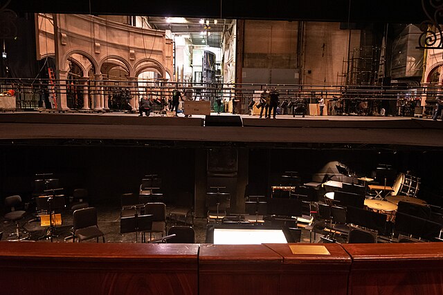 Lyric Opera House Backstage view of a theater showing stage crew at work, set structures, and an orchestra pit with empty music stands and chairs in the foreground.