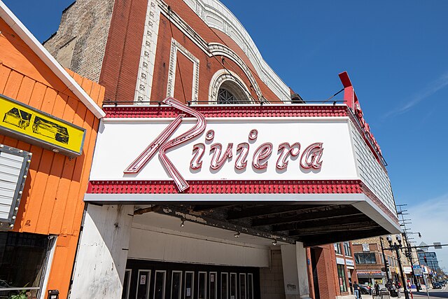 Riviera_Theatre_Chicago The marquee of the Riviera Theatre in Chicago, featuring large red script lettering and bordered lights, is shown against a blue sky and surrounding buildings.