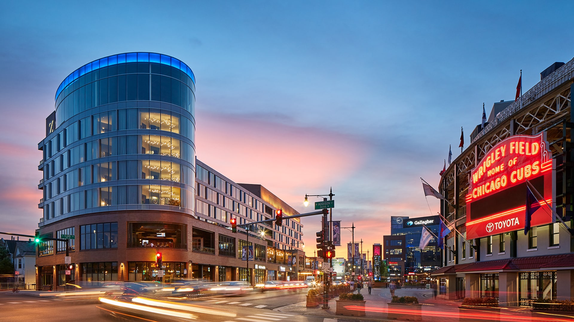 Modern glass building and light trails at dusk near the illuminated marquee of Wrigley Field, home of the Chicago Cubs, in Chicago.