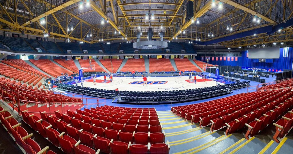 Wide view of an empty indoor basketball arena with red seats, a wooden court, and overhead lights. Basketball hoops and benches are set up on the court.