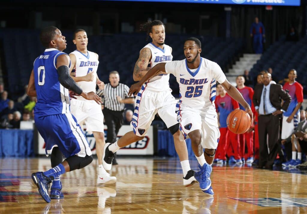 A DePaul basketball player dribbles the ball while being guarded by an opposing player, with teammates and a referee visible in the background.