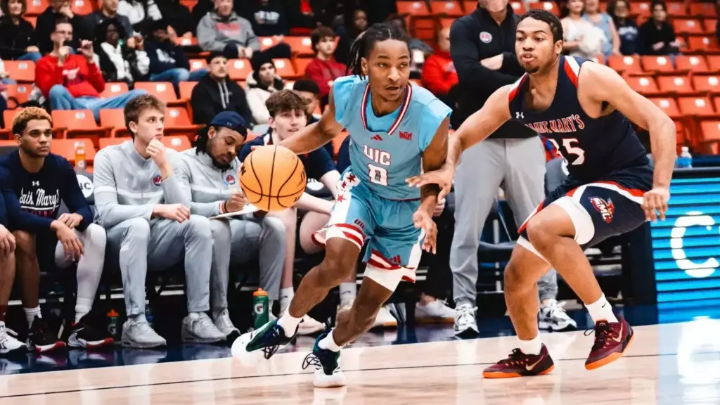 A basketball player in a light blue uniform dribbles the ball while being defended by a player in a dark blue uniform during a game. Spectators are seated in the background.