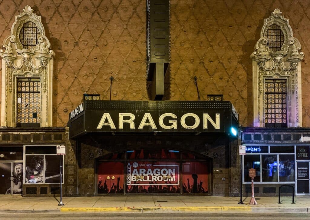 The exterior of the Aragon Ballroom at night, featuring its marquee sign and decorative architecture, with the entrance illuminated and posters visible through the windows.