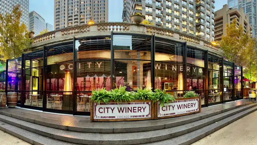 Exterior view of City Winery Chicago, a glass-walled restaurant and event space with tall buildings in the background and greenery in front.