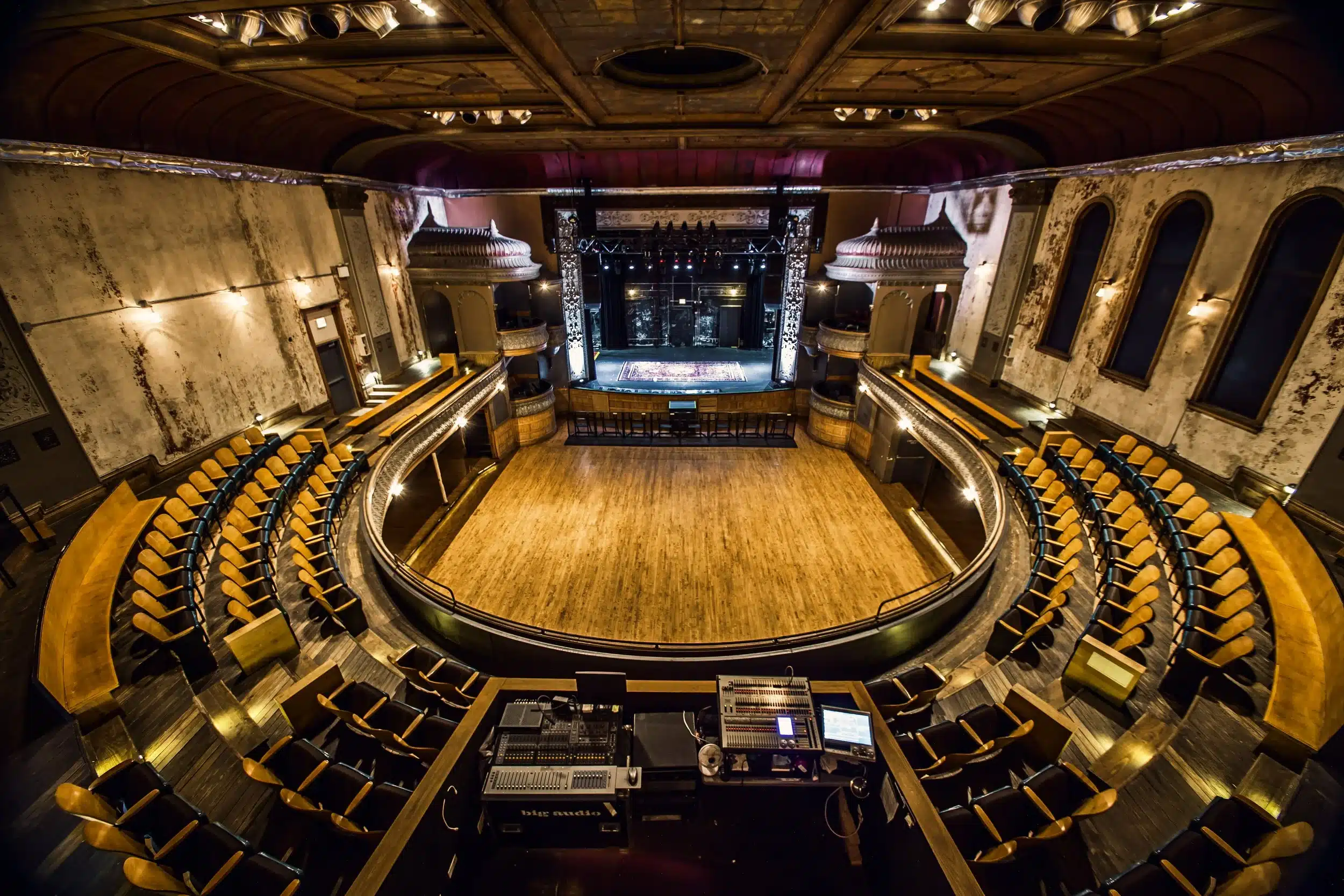 Wide-angle view of an empty historic theater with curved balcony seating, wooden floors, and a lit stage at the far end.