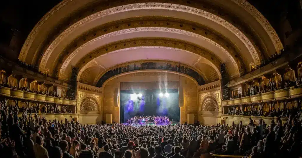 A packed audience watches a live performance on a brightly lit stage inside an ornate, domed theater with multiple balconies.