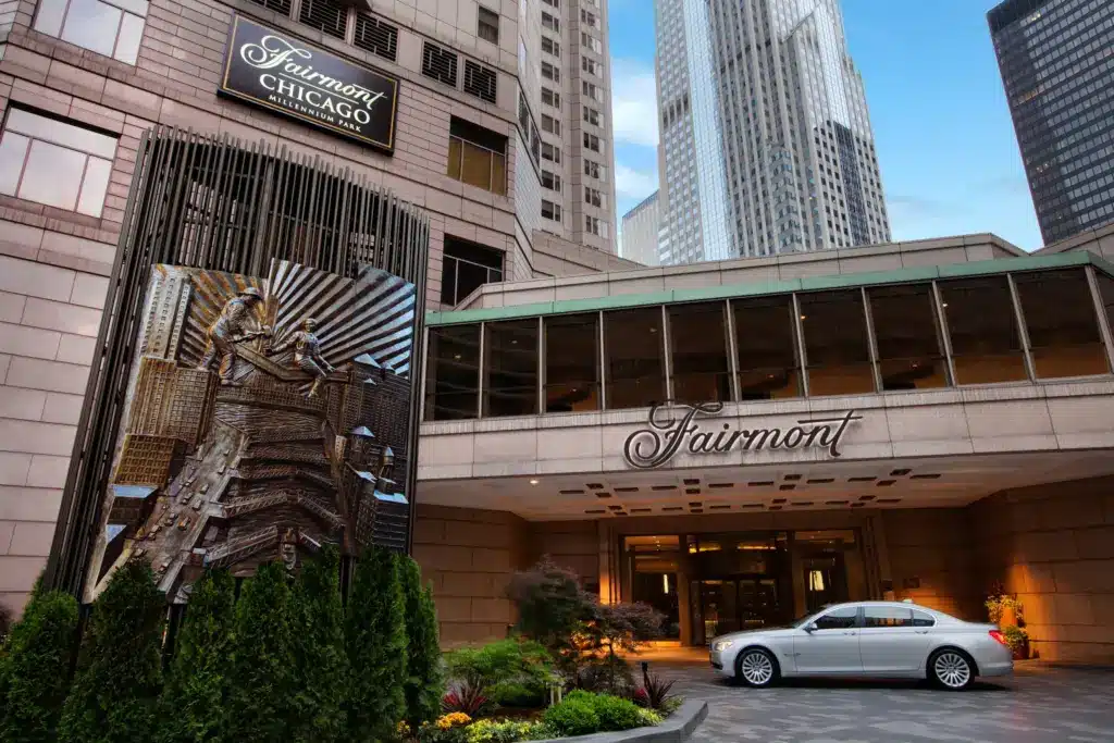 The entrance of the Fairmont Chicago hotel features a large sign, decorative metal sculpture, and a white car parked in front of the main doors on a sunny day.
