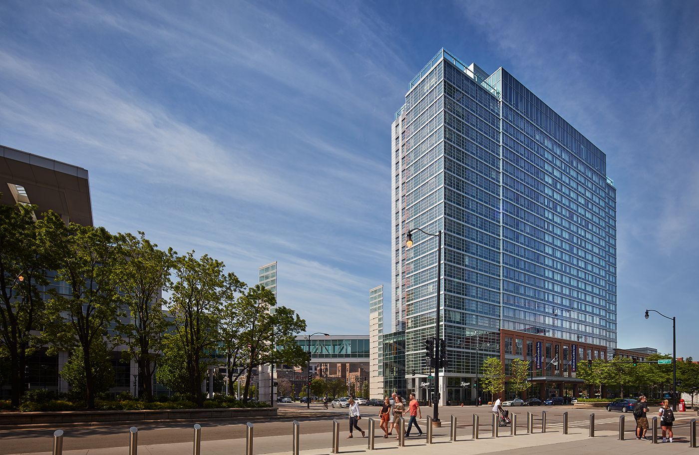 A tall glass office building stands on a city street corner with people walking on the sidewalk and trees lining the road under a clear blue sky.