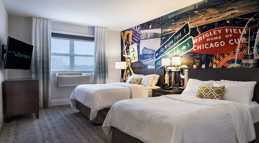 A hotel room with two double beds, a wall mural of Chicago landmarks, a window, an air conditioner, a TV, and bedside lamps.