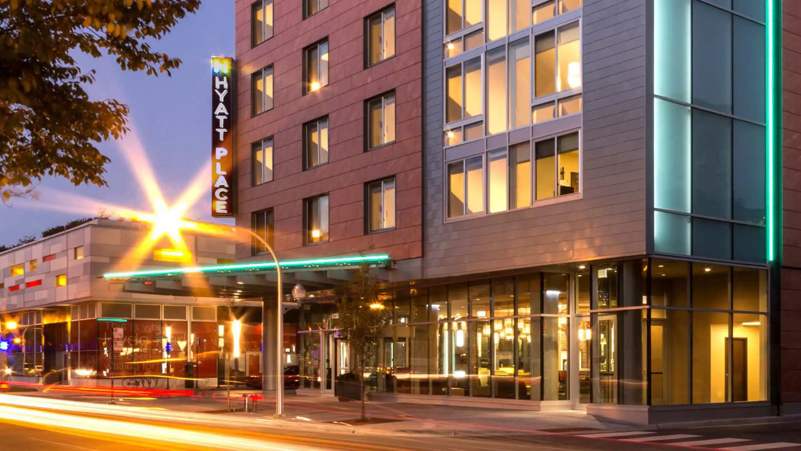 Exterior view of a Hyatt Place hotel building at dusk with lights on inside and light trails from passing cars in the foreground.