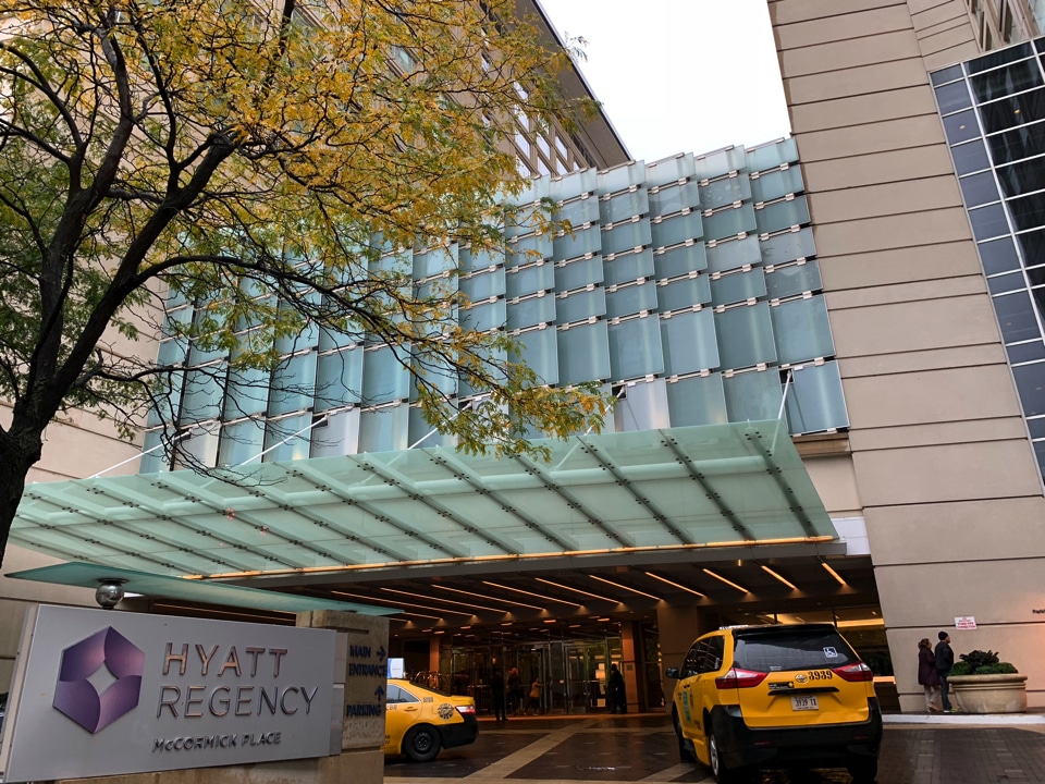 The entrance of the Hyatt Regency McCormick Place hotel, with a glass canopy overhead, several yellow taxis, and a tree with autumn leaves in the foreground.