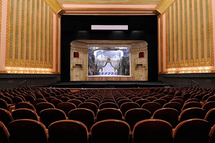 Empty theater with rows of red seats facing an ornate stage set for a performance, featuring gold details and a decorative backdrop.