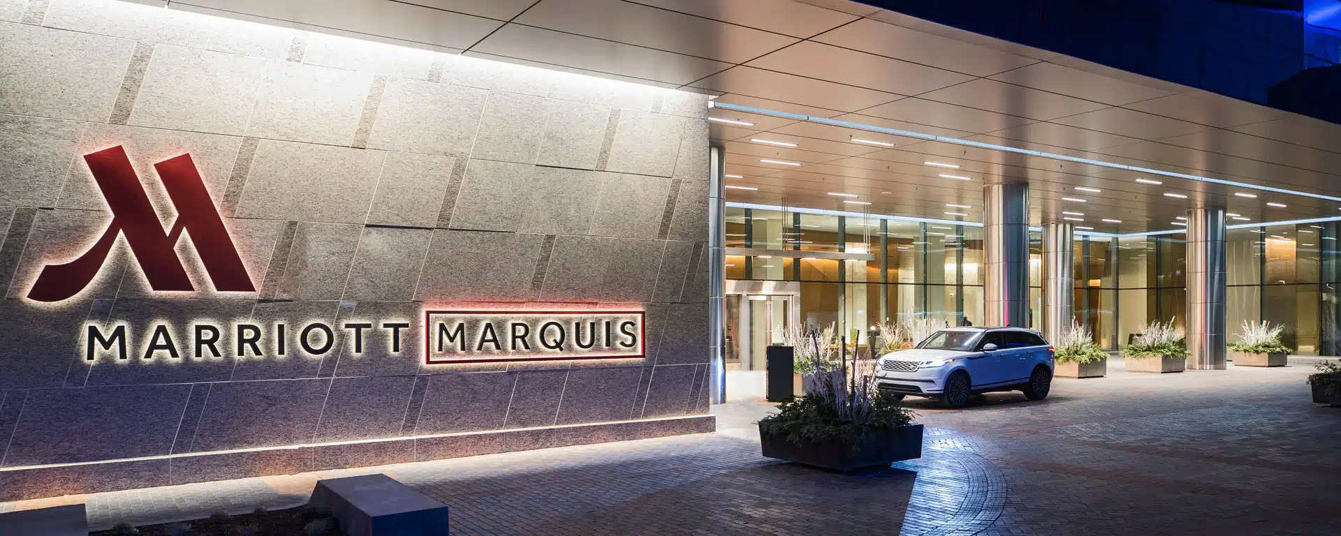 Exterior view of the Marriott Marquis hotel entrance at night, with a white SUV parked near the glass doors and modern landscaping visible.