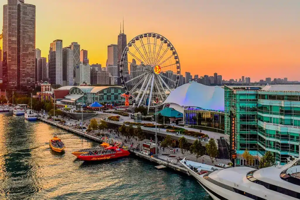 A waterfront cityscape at sunset with a large Ferris wheel, boats docked along the shore, and modern buildings in the background.