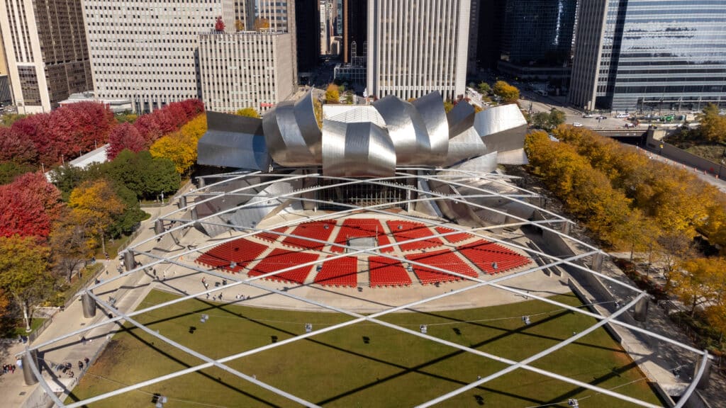 Aerial view of Jay Pritzker Pavilion in Chicago’s Millennium Park, showing red seating, metallic stage structure, and surrounding trees with autumn foliage.