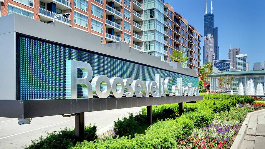 Large sign reading "Roosevelt Collection" in front of modern apartment buildings, with flowers, greenery, and a city skyline in the background.