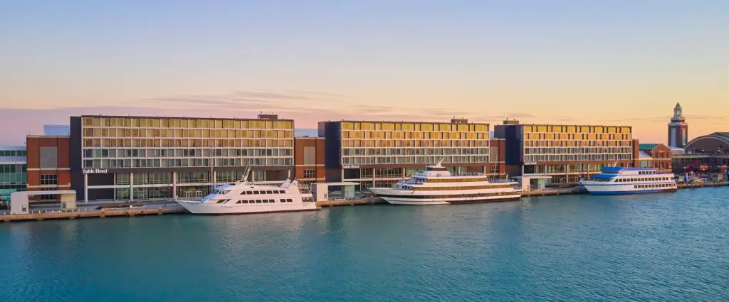 Modern waterfront hotel with large windows and several docked yachts on a calm body of water at sunset, with a distant pier and tower visible.