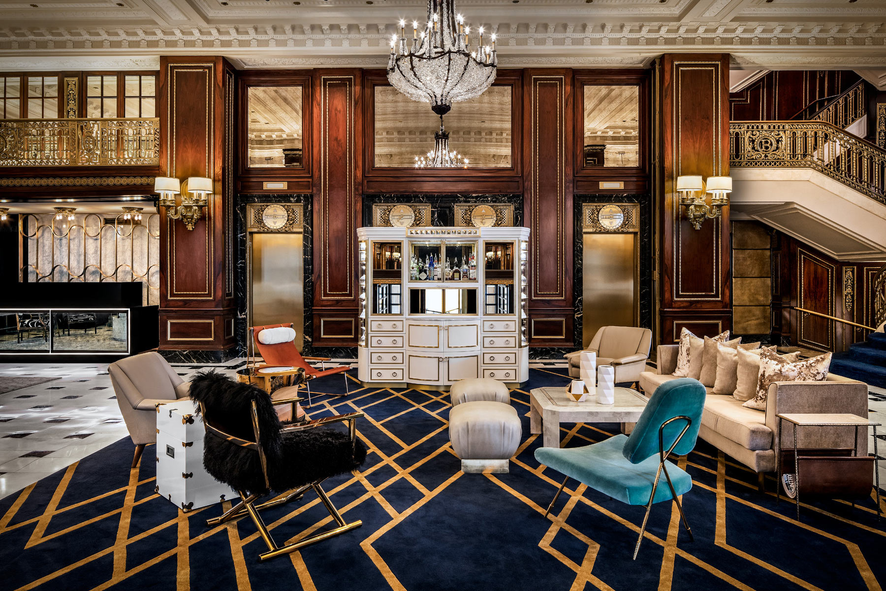 Luxurious hotel lobby with eclectic modern furniture, a central bar cabinet, chandeliers, marble floor, and ornate wood-paneled walls with gold accents and mirrored elevator doors.