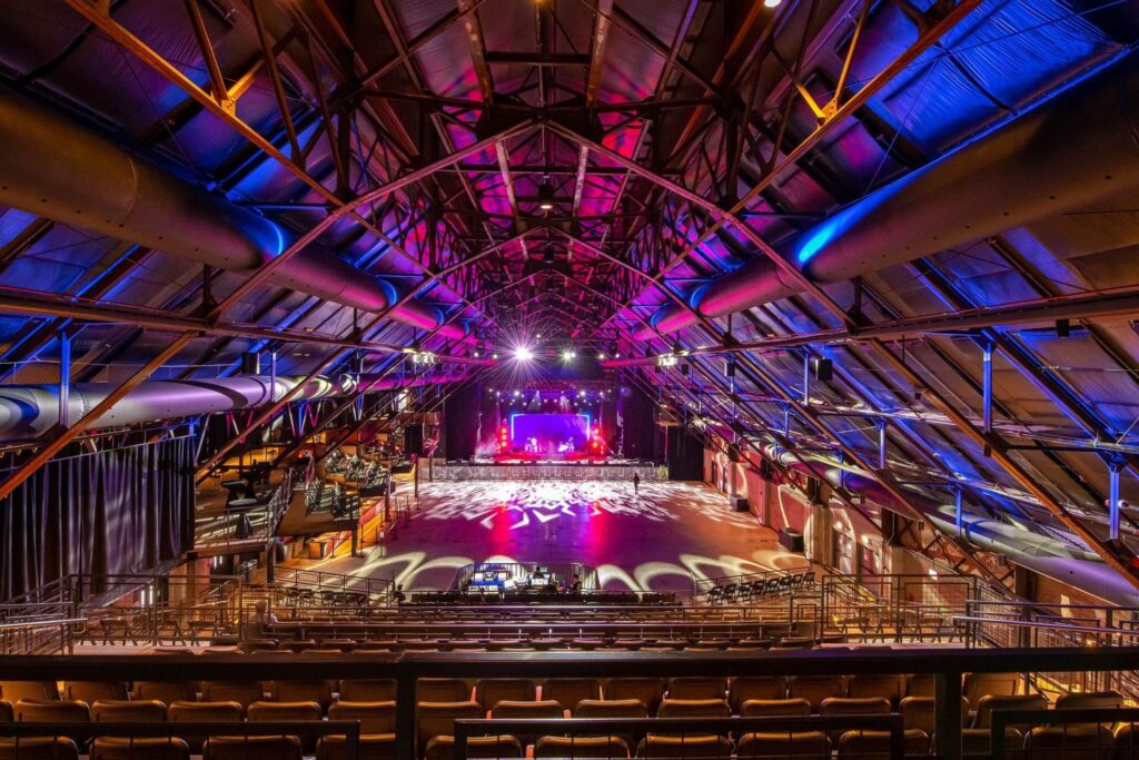 Large indoor concert venue with an empty floor, stage at the far end, colorful lighting, and exposed metal beams in the ceiling.