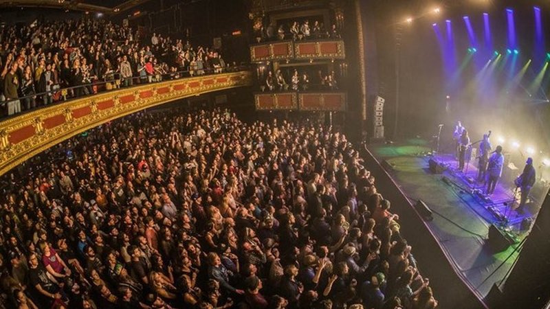 A large audience watches a live band perform on a brightly lit stage in an ornate, crowded theater.