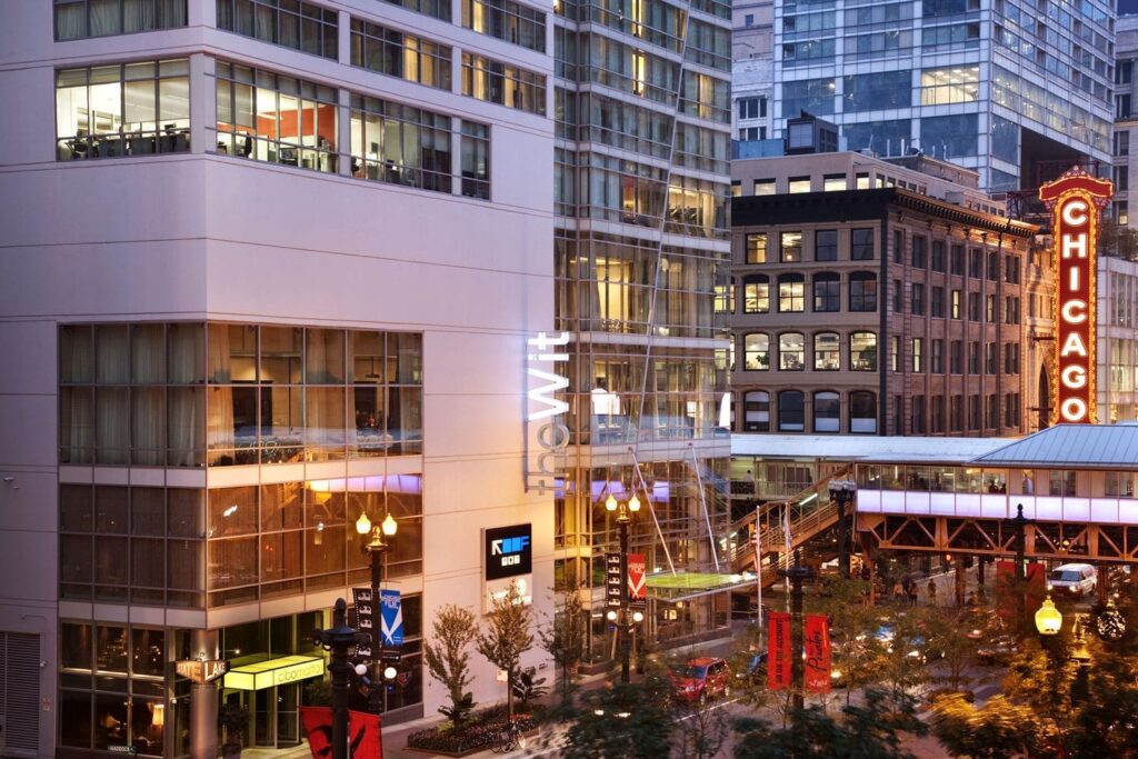 A city street scene featuring The Wit hotel building, the elevated CTA train tracks, and the illuminated Chicago Theatre sign in downtown Chicago at dusk.